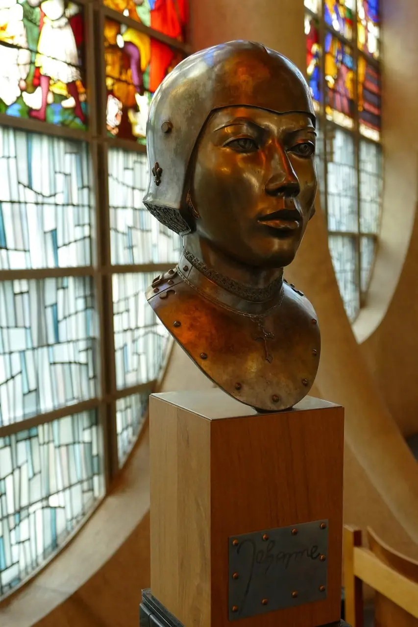 Bronze bust of Joan of Arc in armor, displayed inside a church in Normandy, France, with stained glass in the background