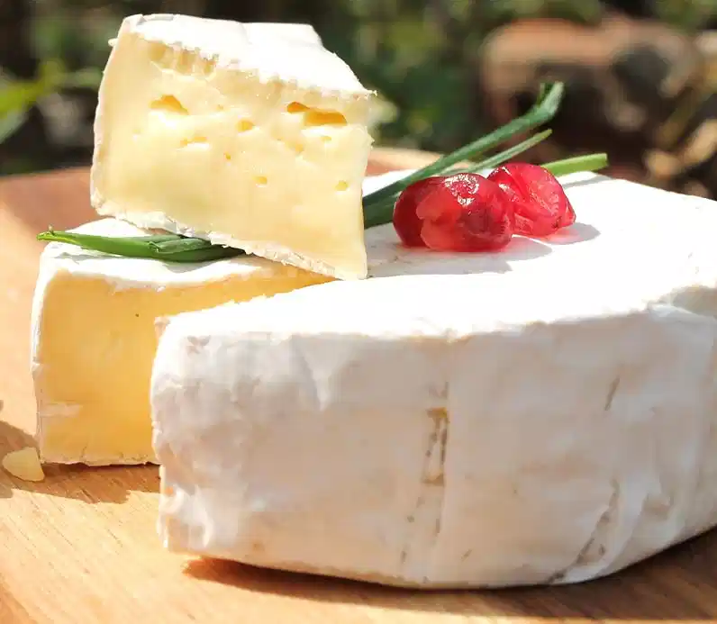 Close-up of creamy Camembert cheese with white rind, garnished with red berries and herbs on a wooden board – Normandy speciality