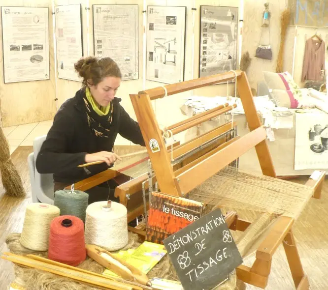 Woman demonstrating hand weaving on a wooden loom with yarns and textile samples – traditional Norman craft
